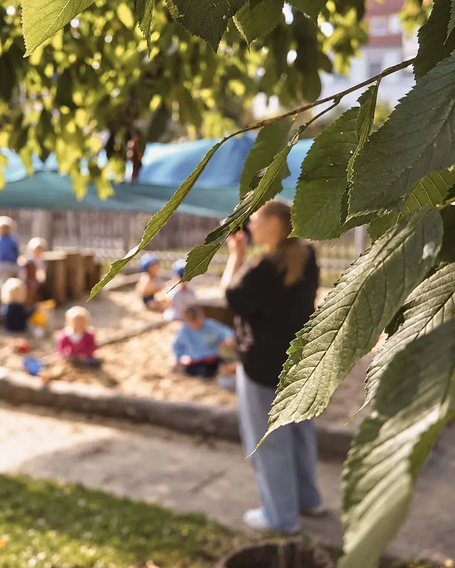 Blick durch Baumblätter auf eine Fotografin im Garten, davor spielen Kinder unscharf im Sand