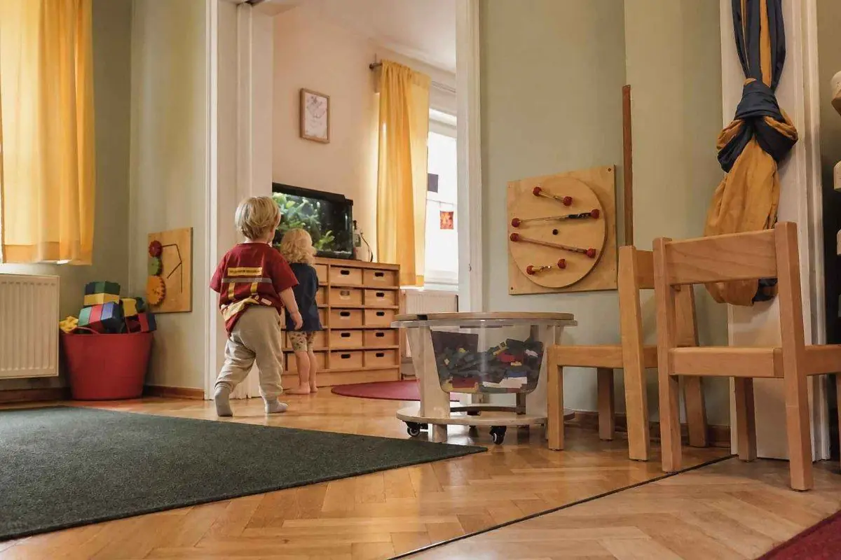 Gruppenraum mit Holzmöbeln, in dem Kinder und eine Erzieherin spielen