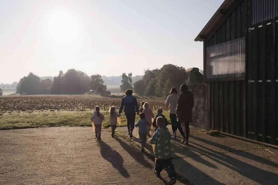 Kinder und Begleitpersonen gehen im Morgenlicht an einer Scheune vorbei über den Hof