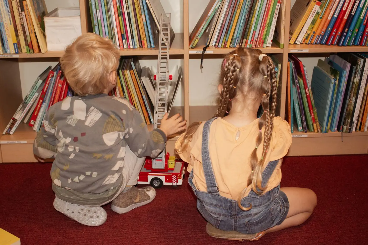 Zwei Kinder sitzen von hinten vor einem vollen Bücherregal in der Bücherei