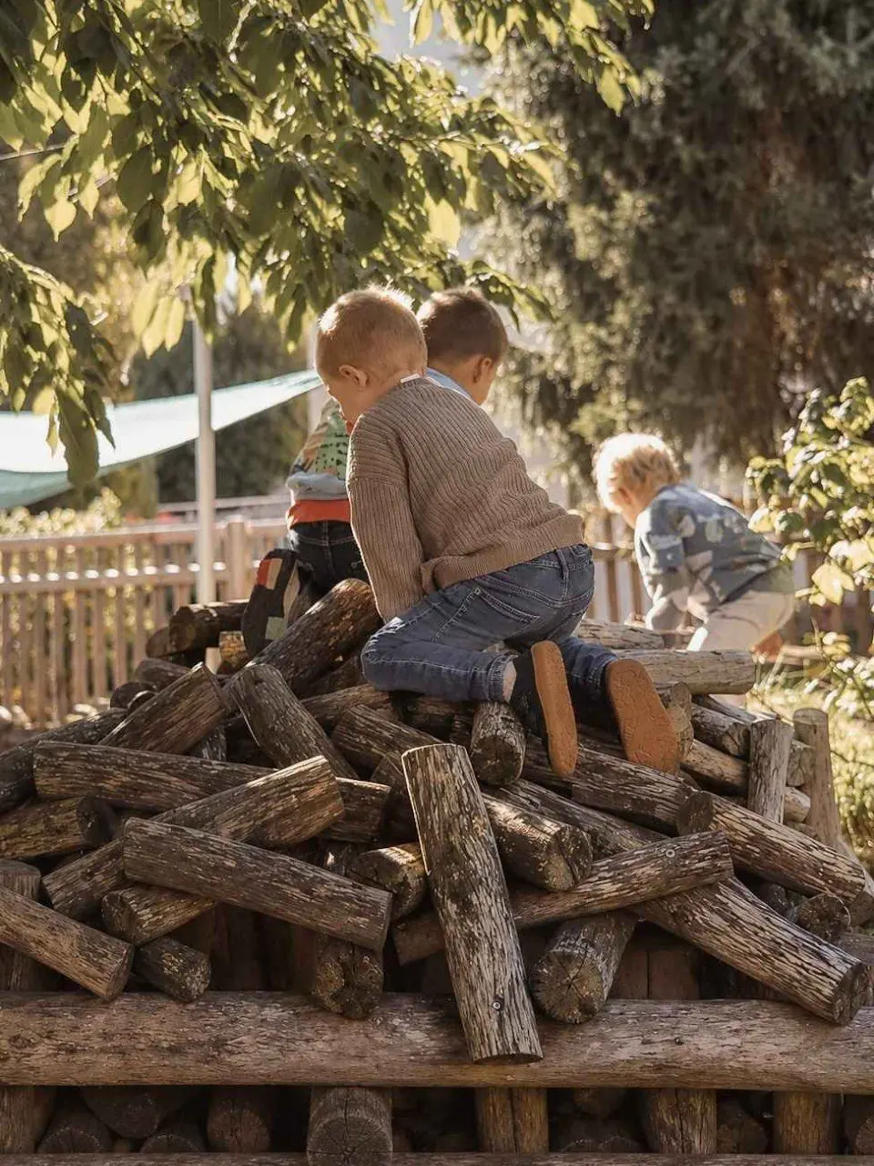 Kinder klettern auf einem Stapel aus Rundhölzern unter Bäumen