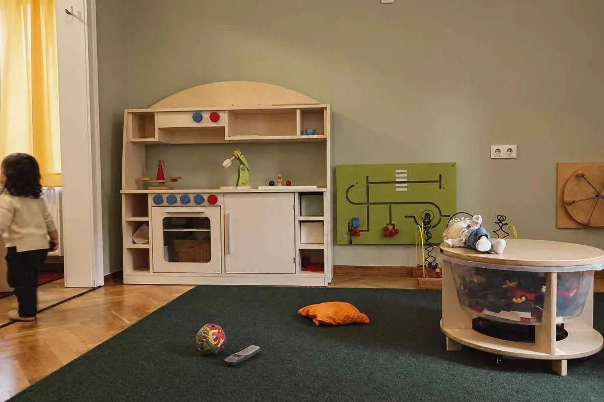 Heller Raum mit Spielküche aus Holz, dunkelgrünem Teppich und einem Kind im Hintergrund