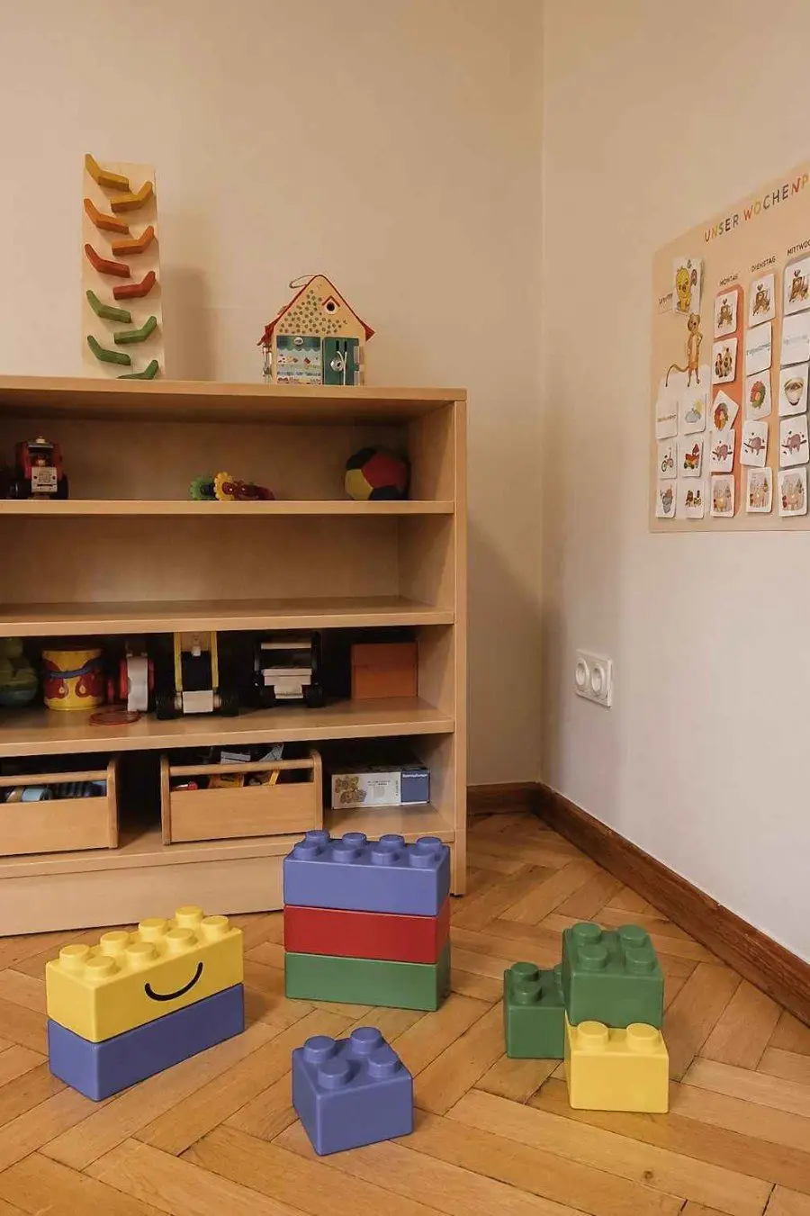 Holzregal mit Spielmaterial, davor große bunte Bausteine, an der Wand eine Bildtafel